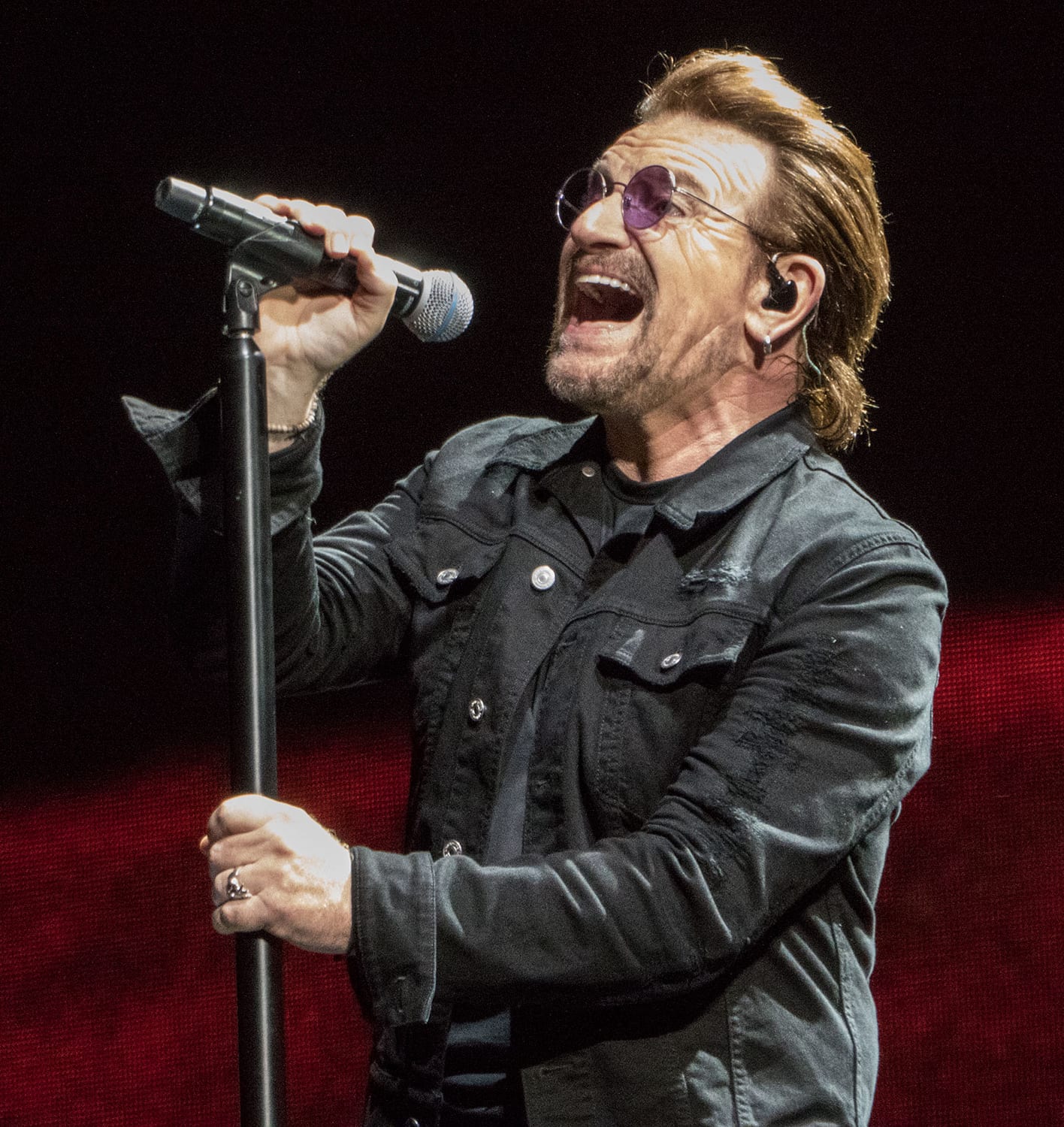 Un hombre con gafas oscuras canta frente a un micrófono bajo luces de escenario cálidas.