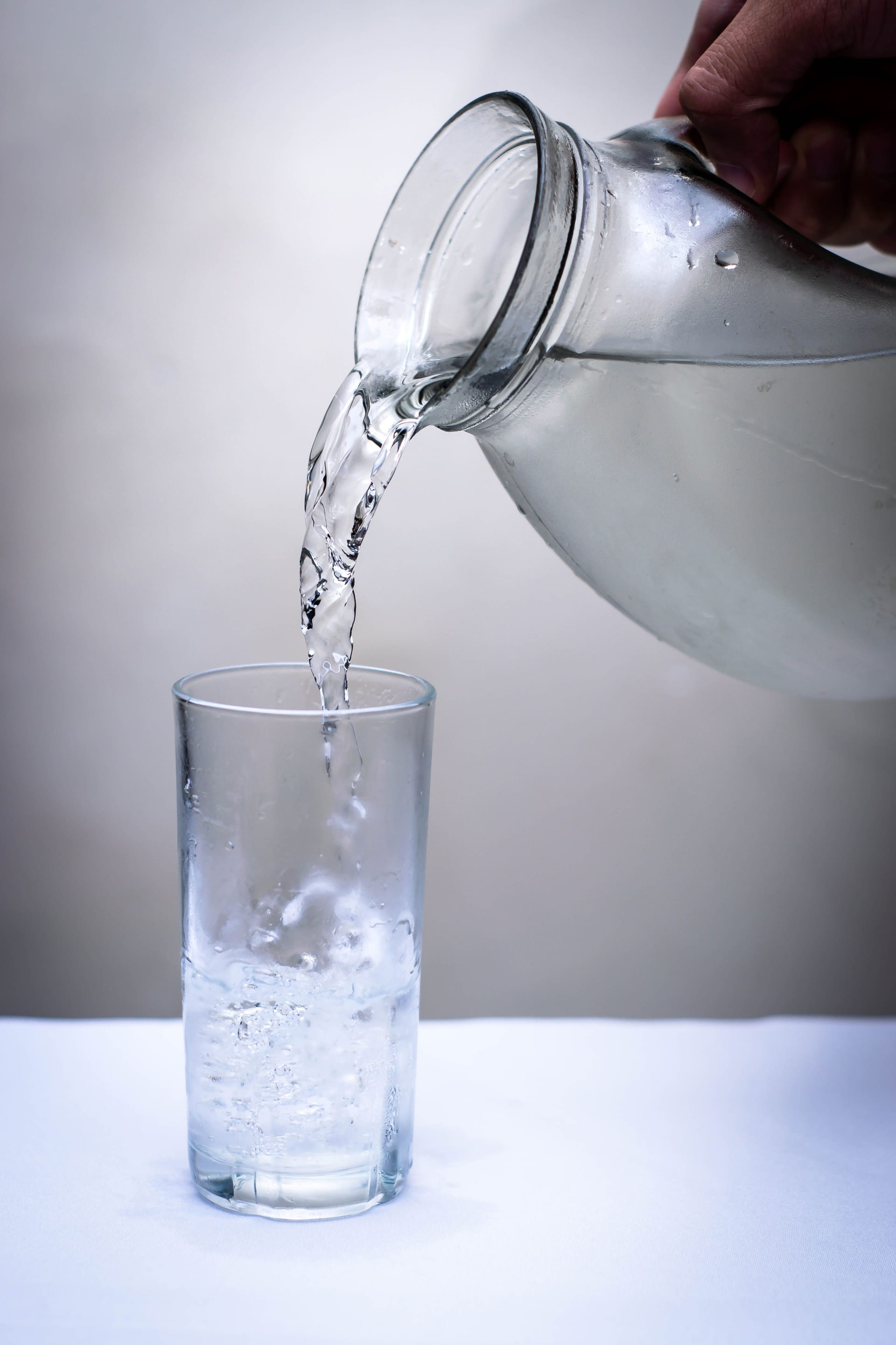 Un chorro cae de una jarra transparente hacia un vaso alto, con el fondo desenfocado en gris claro.