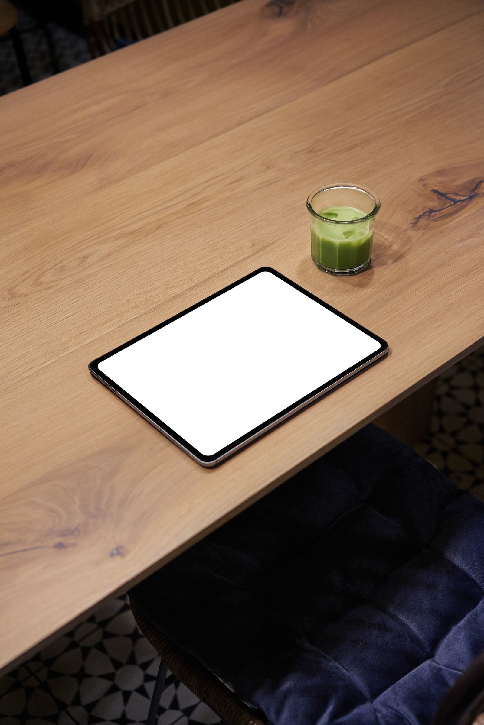 Una tableta con la pantalla en blanco descansa sobre una mesa de madera junto a un vaso con una bebida verde.