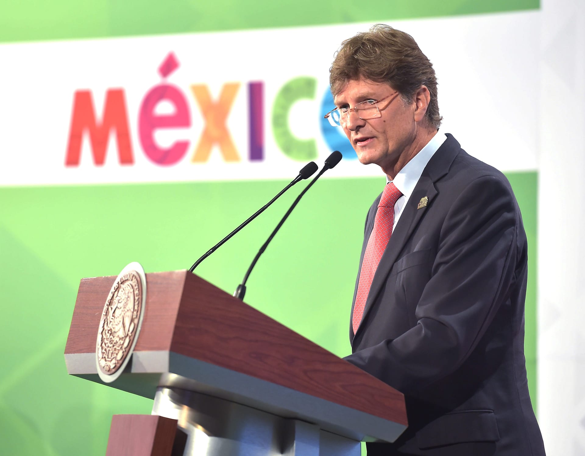 Un hombre con gafas y traje azul marino habla ante un micrófono en un podio con el logotipo de México al fondo.