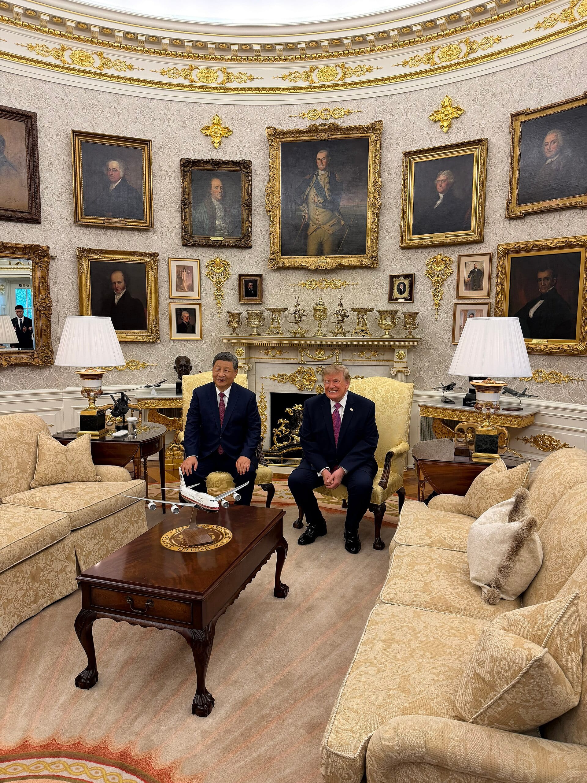 Xi Jinping y Donald Trump sonríen sentados uno junto al otro en el Despacho Oval, con una réplica de avión sobre la mesa frente a ellos.