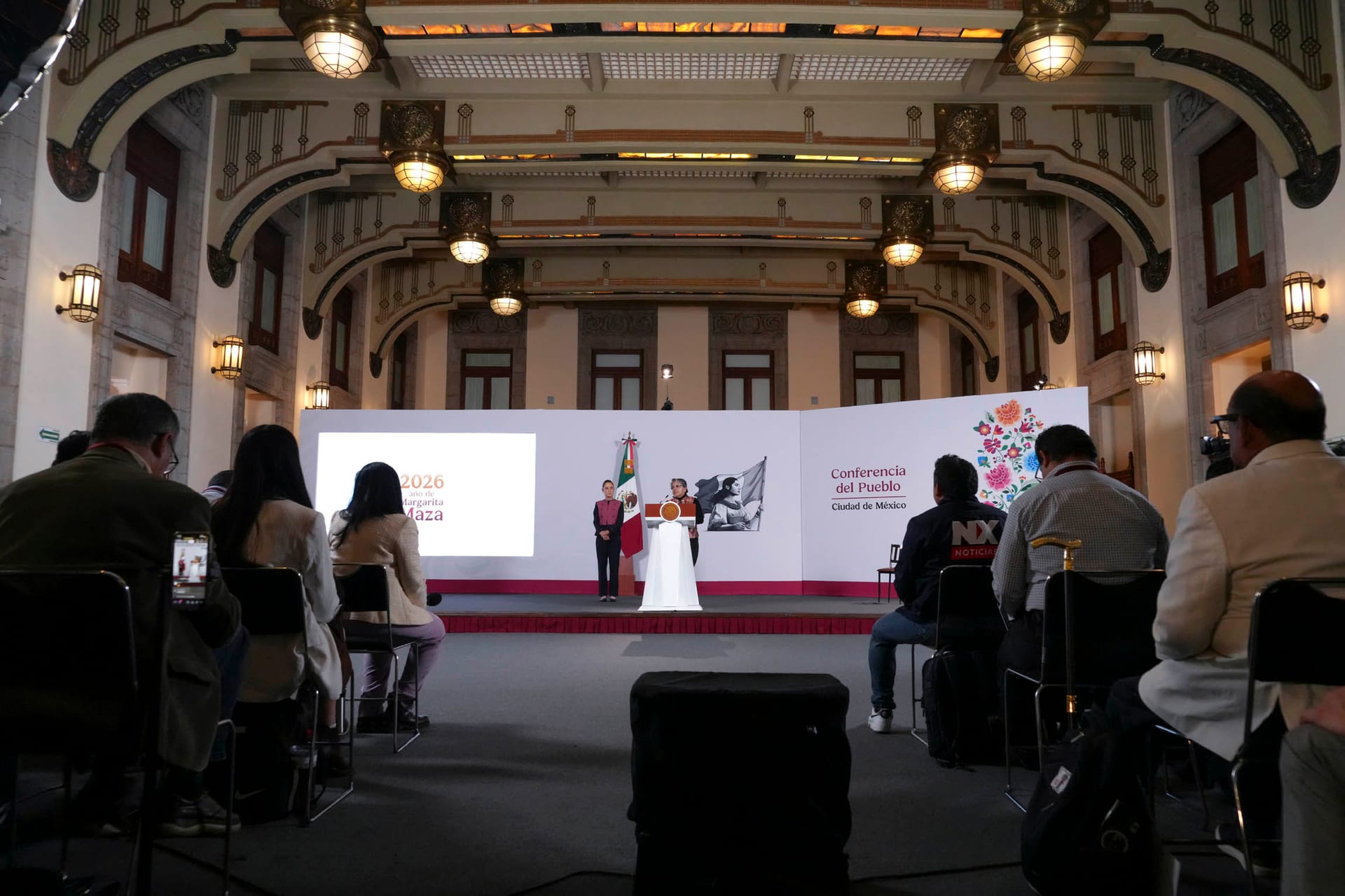 Periodistas sentados de espaldas observan a una oradora en un podio, en un salón con lámparas de estilo art decó.