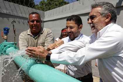 Tres hombres sostienen juntos una llave para abrir una válvula de tubería verde mientras el agua brota a chorros.