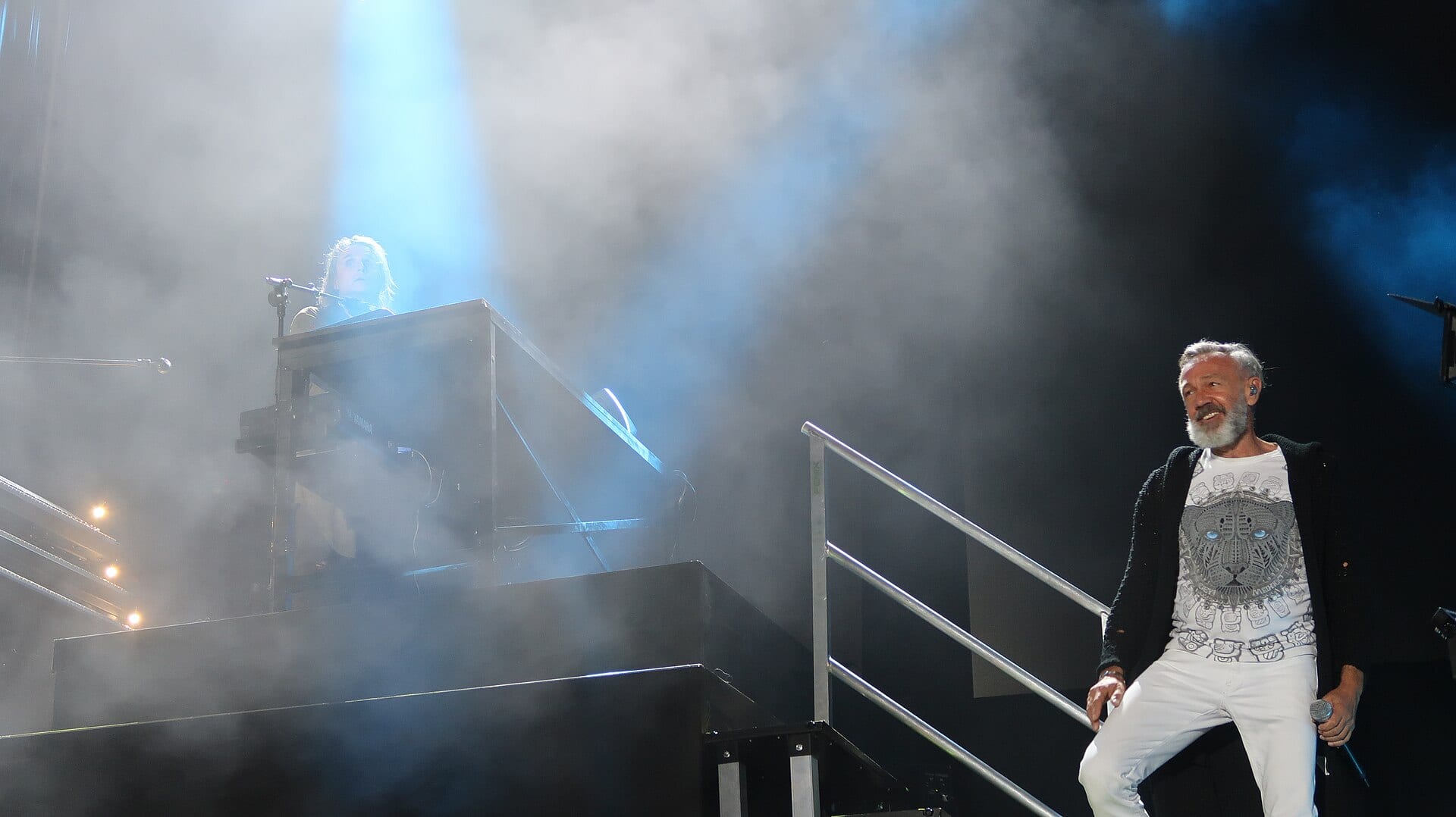 Nacho Cano baja unas escaleras con micrófono en la mano durante un concierto, con un tecladista al fondo entre humo escénico.