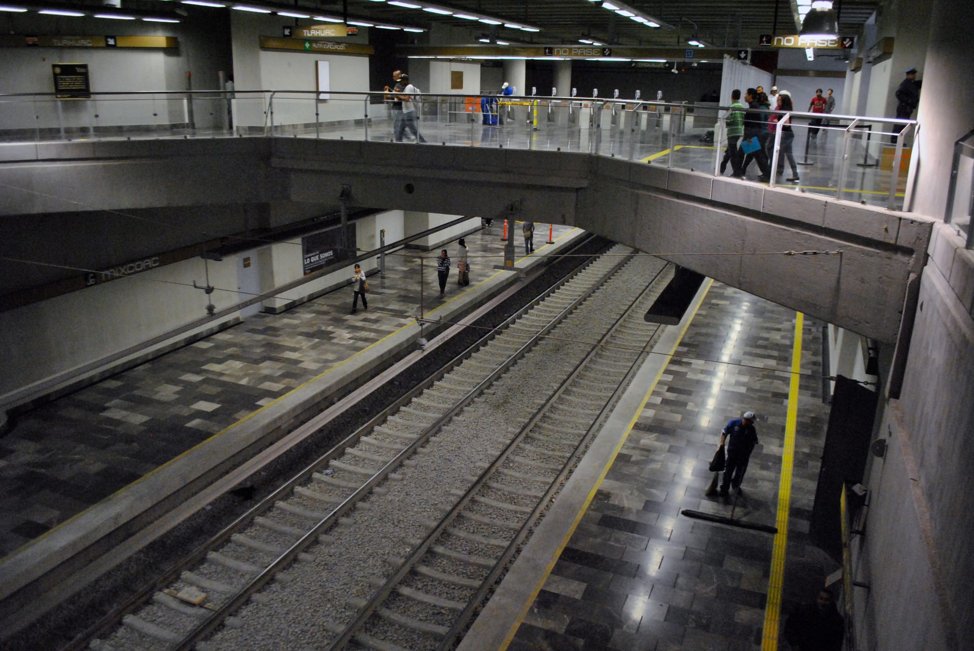 Un andén de una estación del Metro con vías vacías, viajeros en la pasarela superior y un trabajador de limpieza junto a las vías.