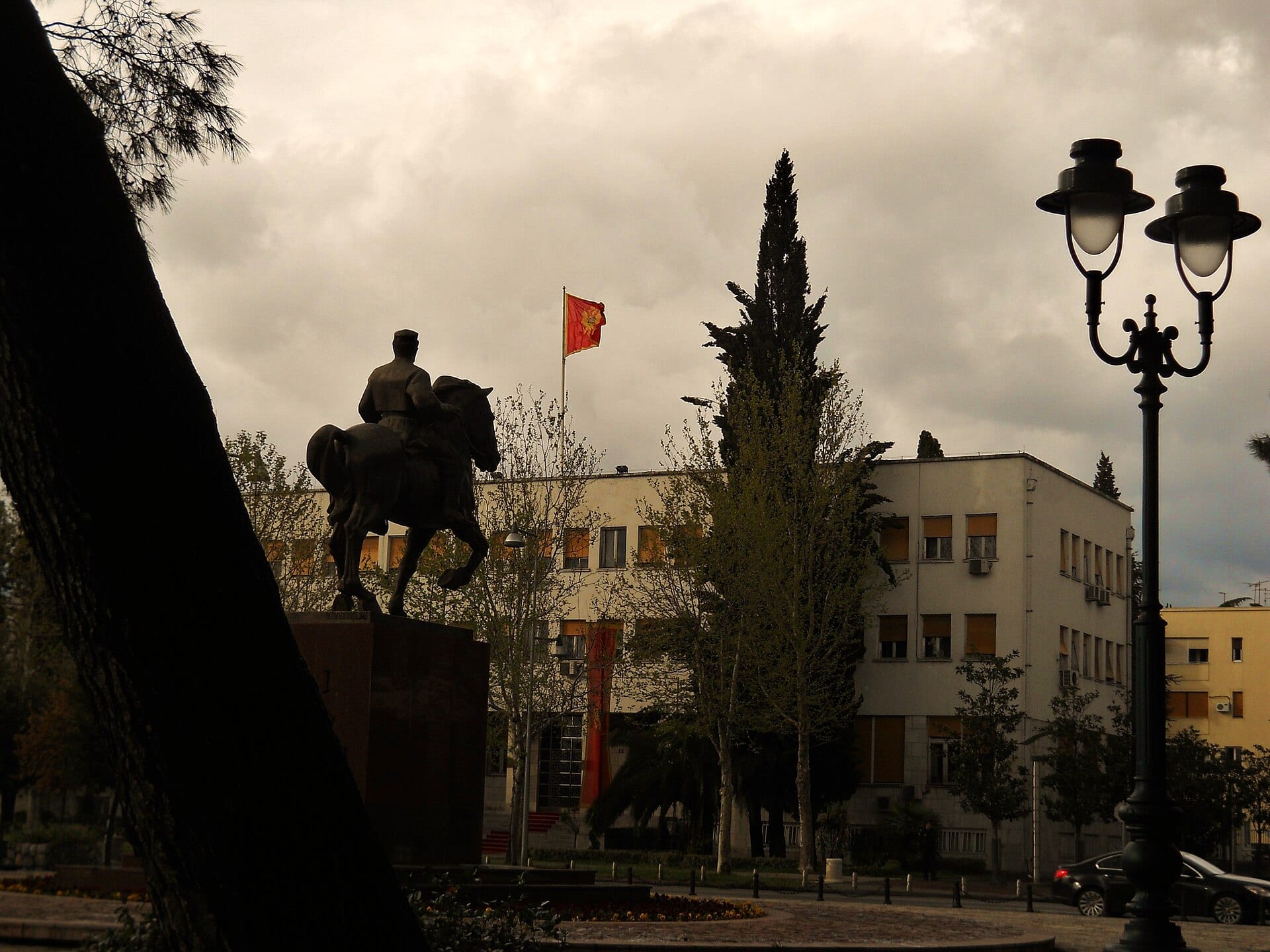 Una estatua ecuestre y la bandera de Montenegro ondeando frente a un edificio de gobierno en Podgorica, bajo un cielo nublado.