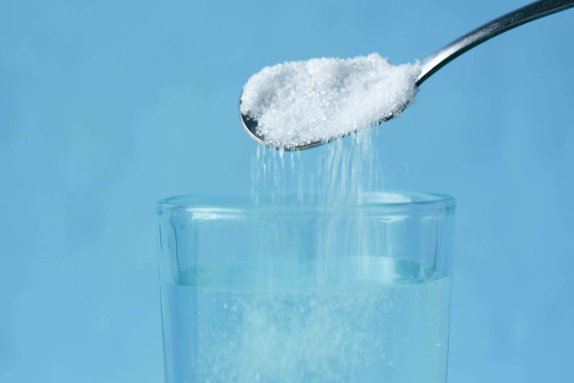 Una cuchara vierte azúcar blanca sobre un vaso con agua, con un fondo azul liso.