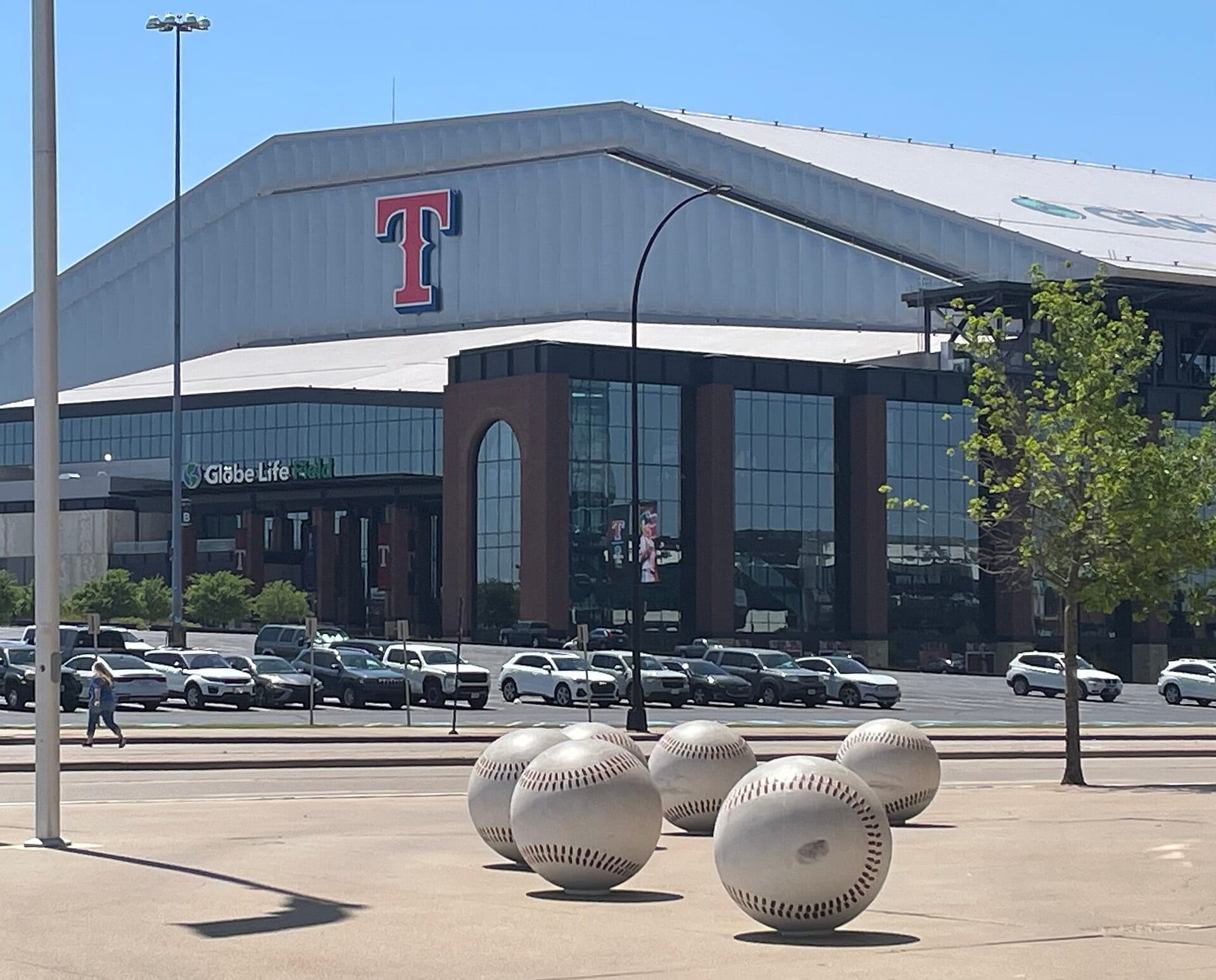 Fachada de Globe Life Field con esculturas de pelotas de beisbol gigantes en el estacionamiento, bajo un cielo despejado.