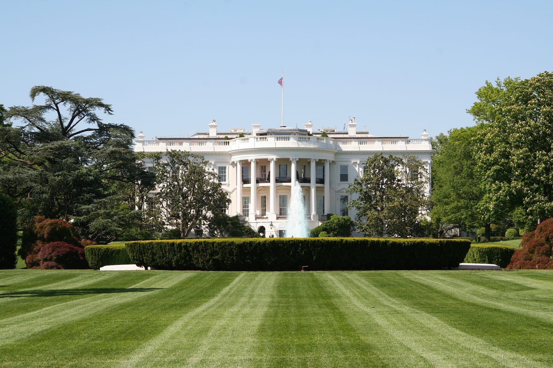 La fachada sur de la Casa Blanca con una fuente activa y un jardín extenso en primer plano, bajo un cielo despejado.
