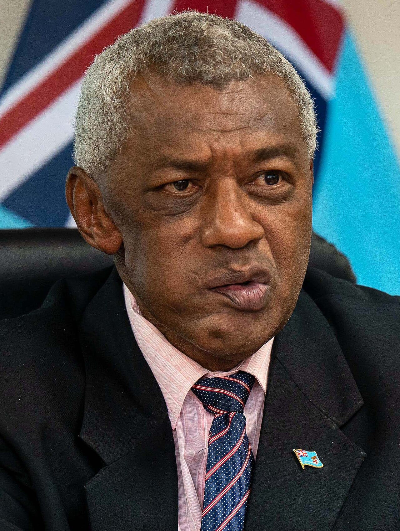 El ministro fiyiano Pio Tikoduadua mira serio hacia la cámara, con las banderas de Fiyi y el Reino Unido desenfocadas detrás.