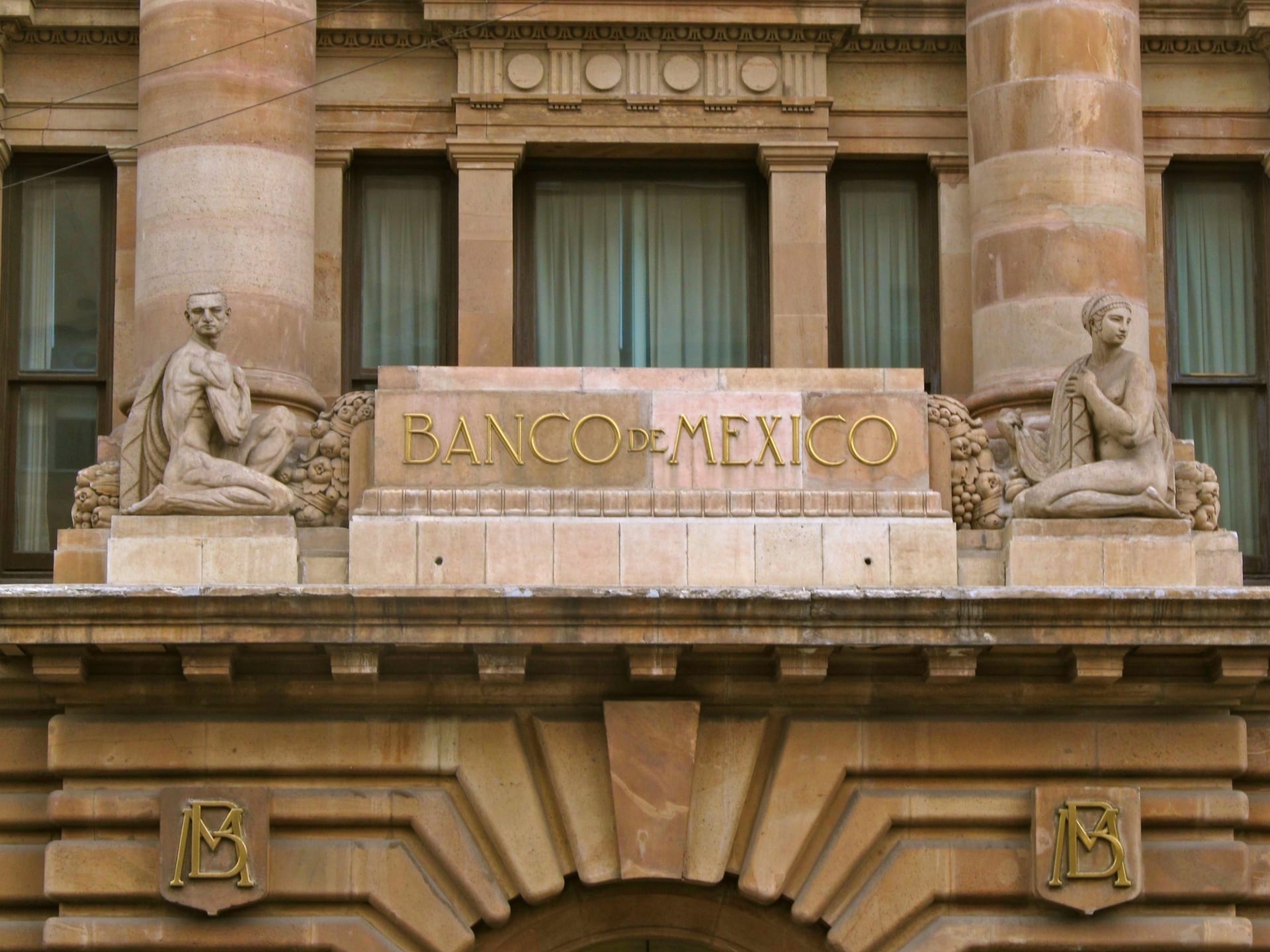 Fachada de piedra de un edificio con el nombre Banco de México en letras doradas, entre dos columnas y dos figuras esculpidas.