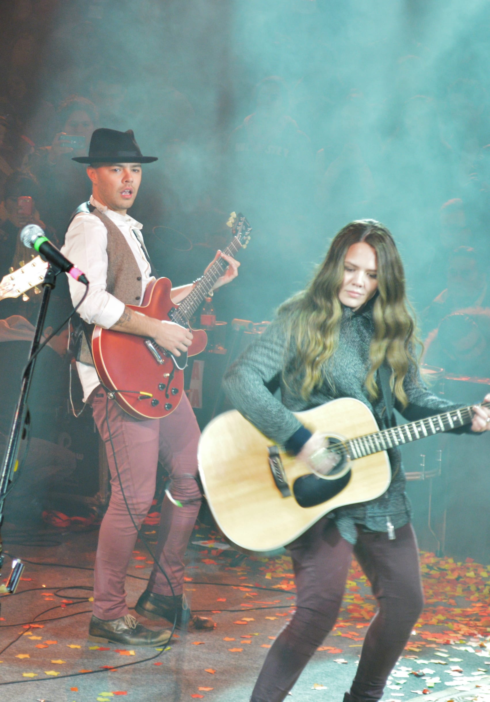 Jesse y Joy Huerta tocan la guitarra juntos sobre un escenario cubierto de confeti, con niebla escénica al fondo.