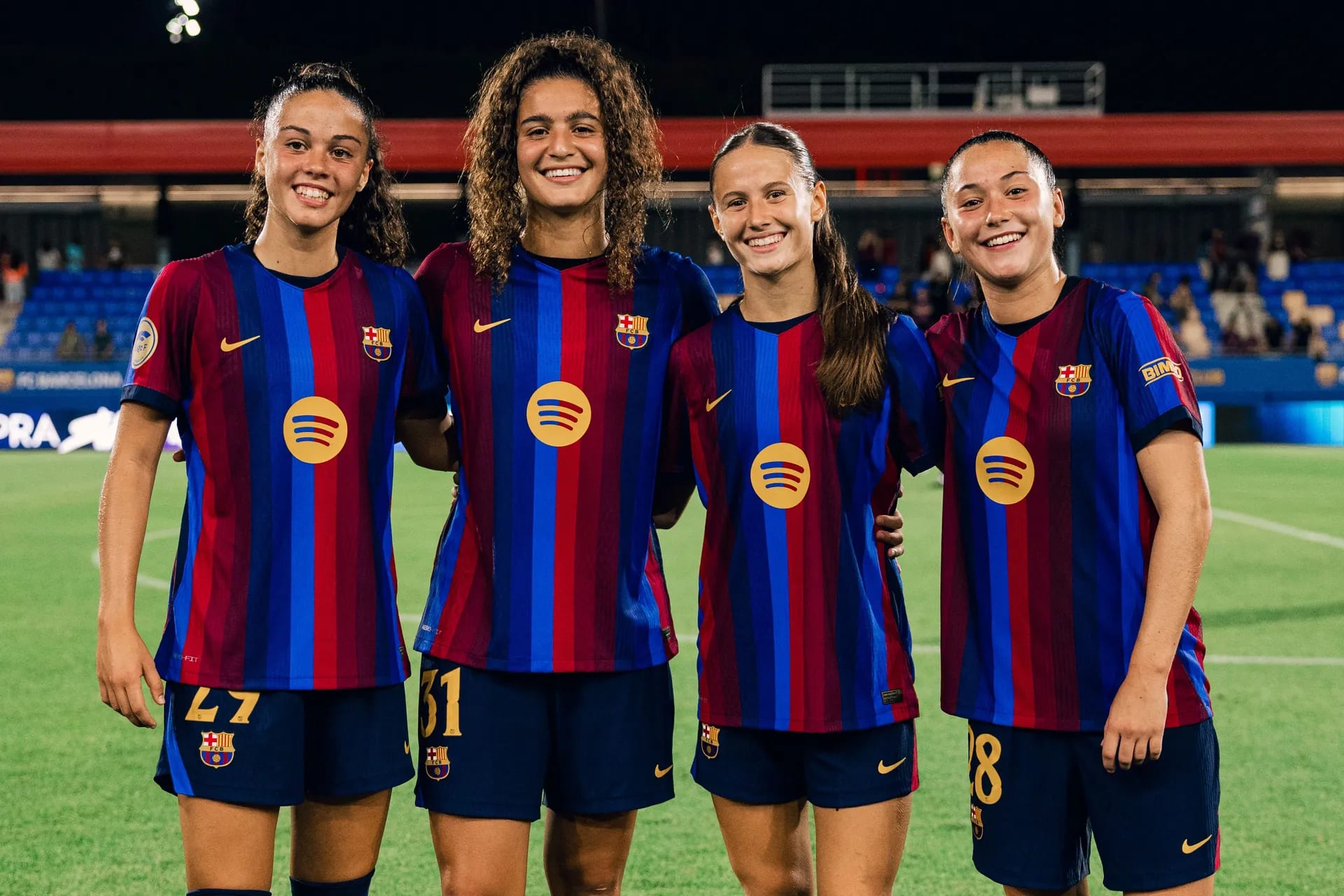 Cuatro futbolistas del Barcelona posan sonrientes y abrazadas sobre el césped tras un partido, con las gradas del estadio al fondo.