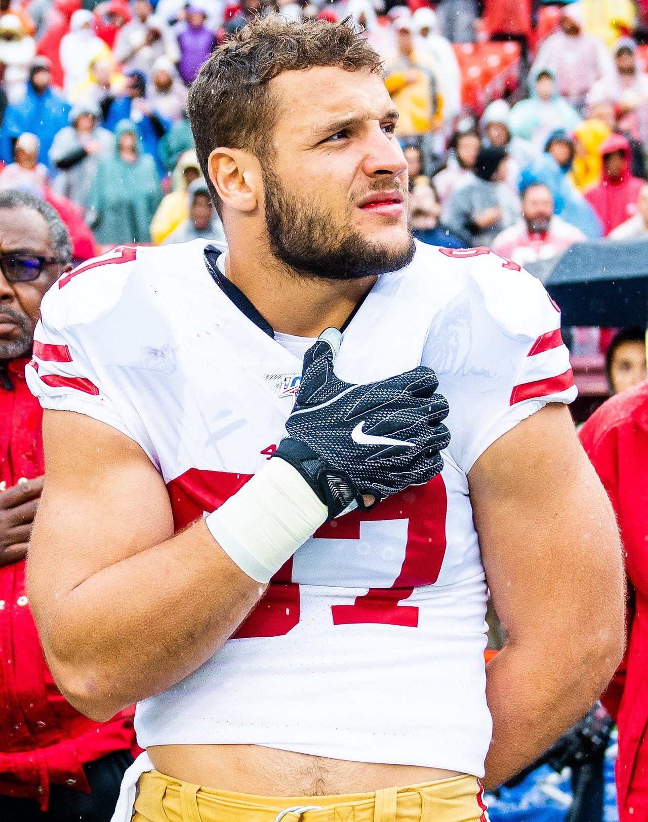 Retrato de un jugador de futbol americano con uniforme blanco y rojo de los 49ers, de pie frente a la grada.