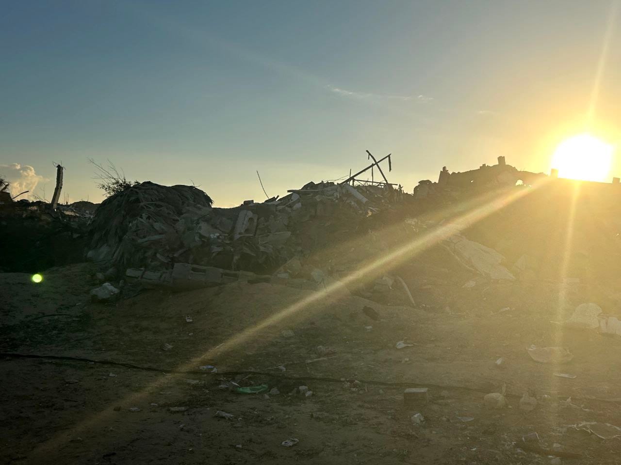 Restos de un edificio derrumbado bajo un cielo con el sol poniéndose al fondo, en un terreno cubierto de escombros.