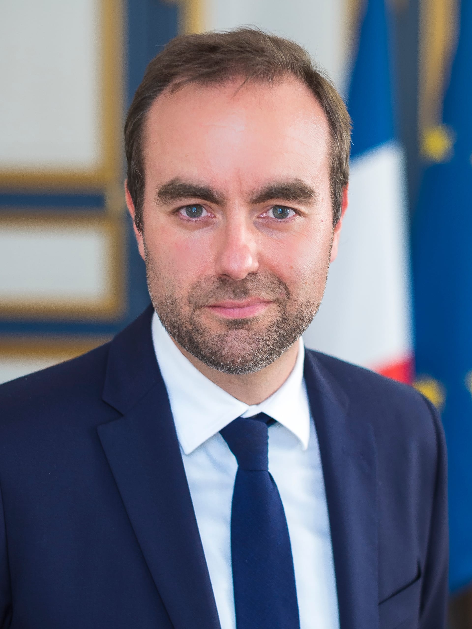 Retrato de un hombre de mediana edad con traje oscuro y corbata azul marino, de pie frente a la bandera de Francia.