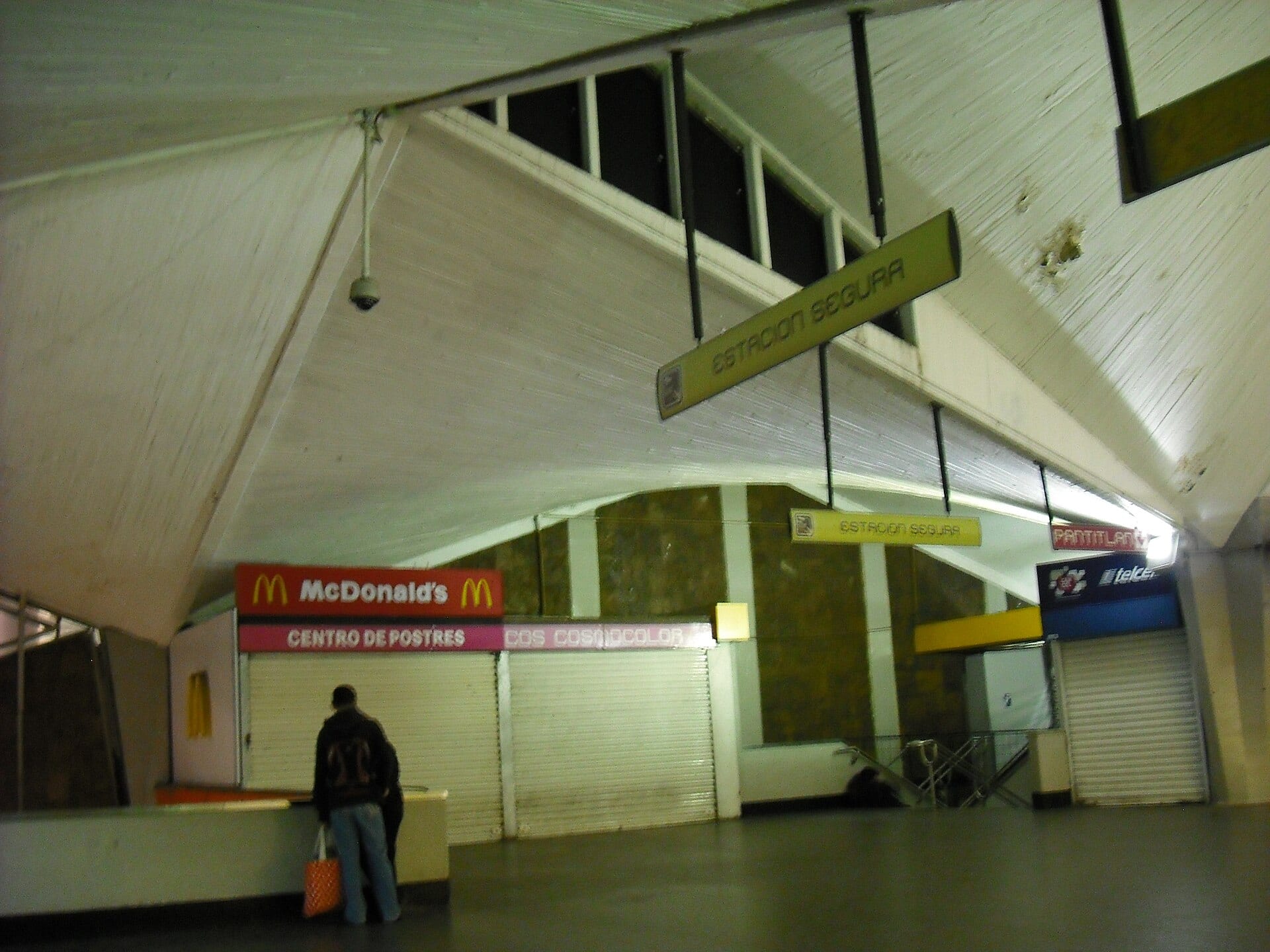El interior techado de una estación del Metro, con locales comerciales, letreros colgantes de "Estación segura" y una persona de espaldas junto a un mostrador.