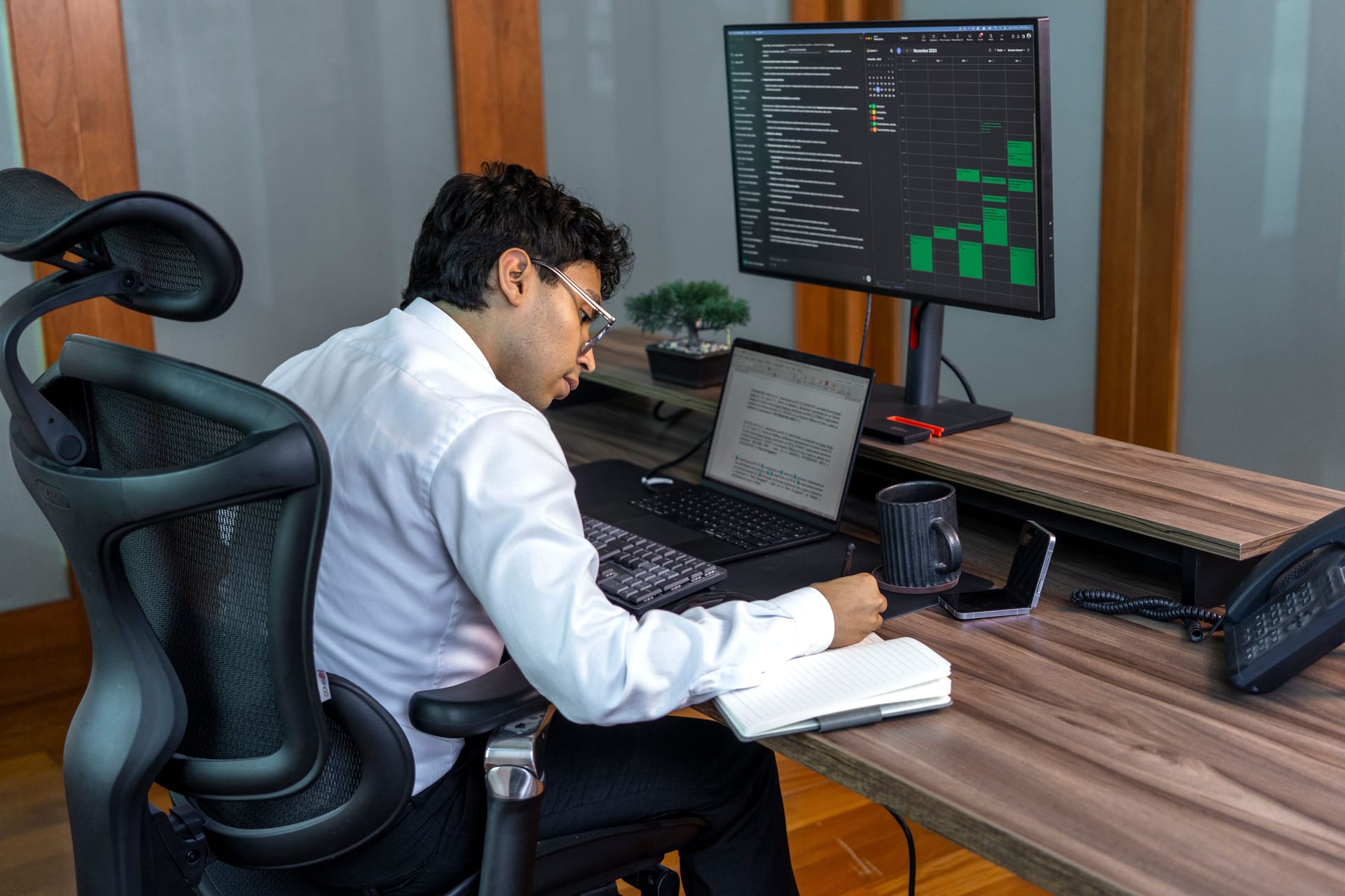 Un hombre con lentes y camisa blanca se inclina hacia adelante frente a una laptop, con un monitor grande y una libreta sobre el escritorio.