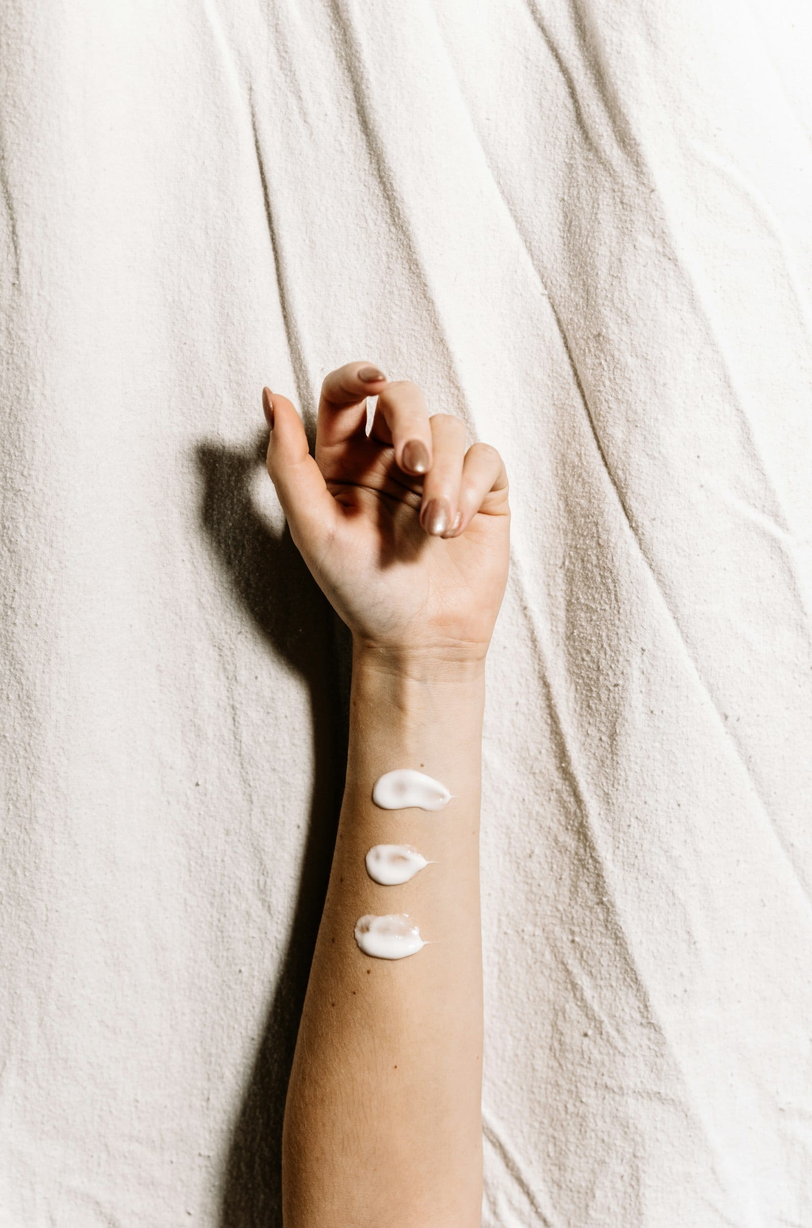 Tres gotas de crema blanca puestas en fila sobre la piel de un antebrazo, sobre una tela clara.