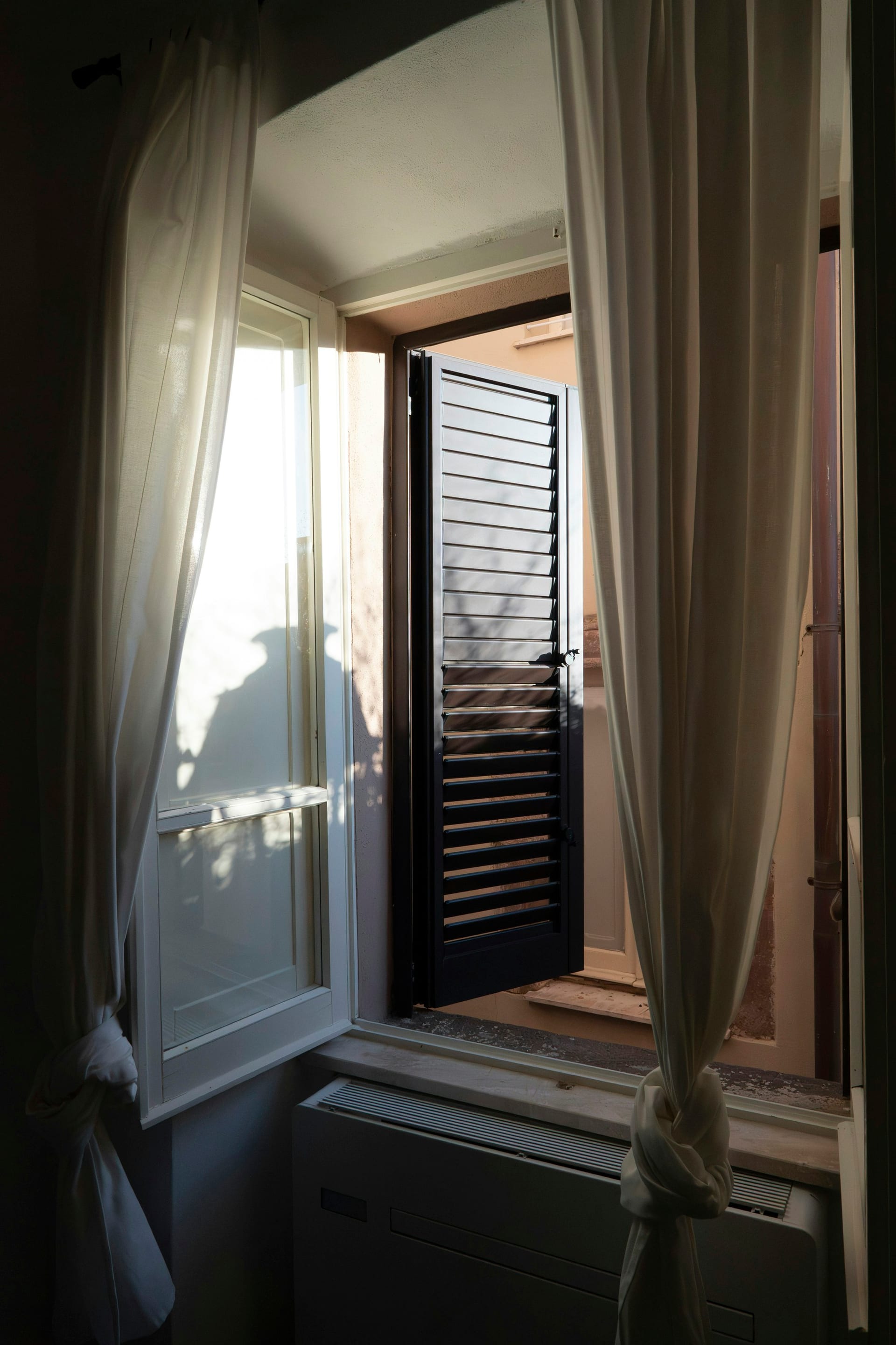 Luz solar de la mañana entra por una ventana abierta con cortinas, proyectando sombras en un cuarto.