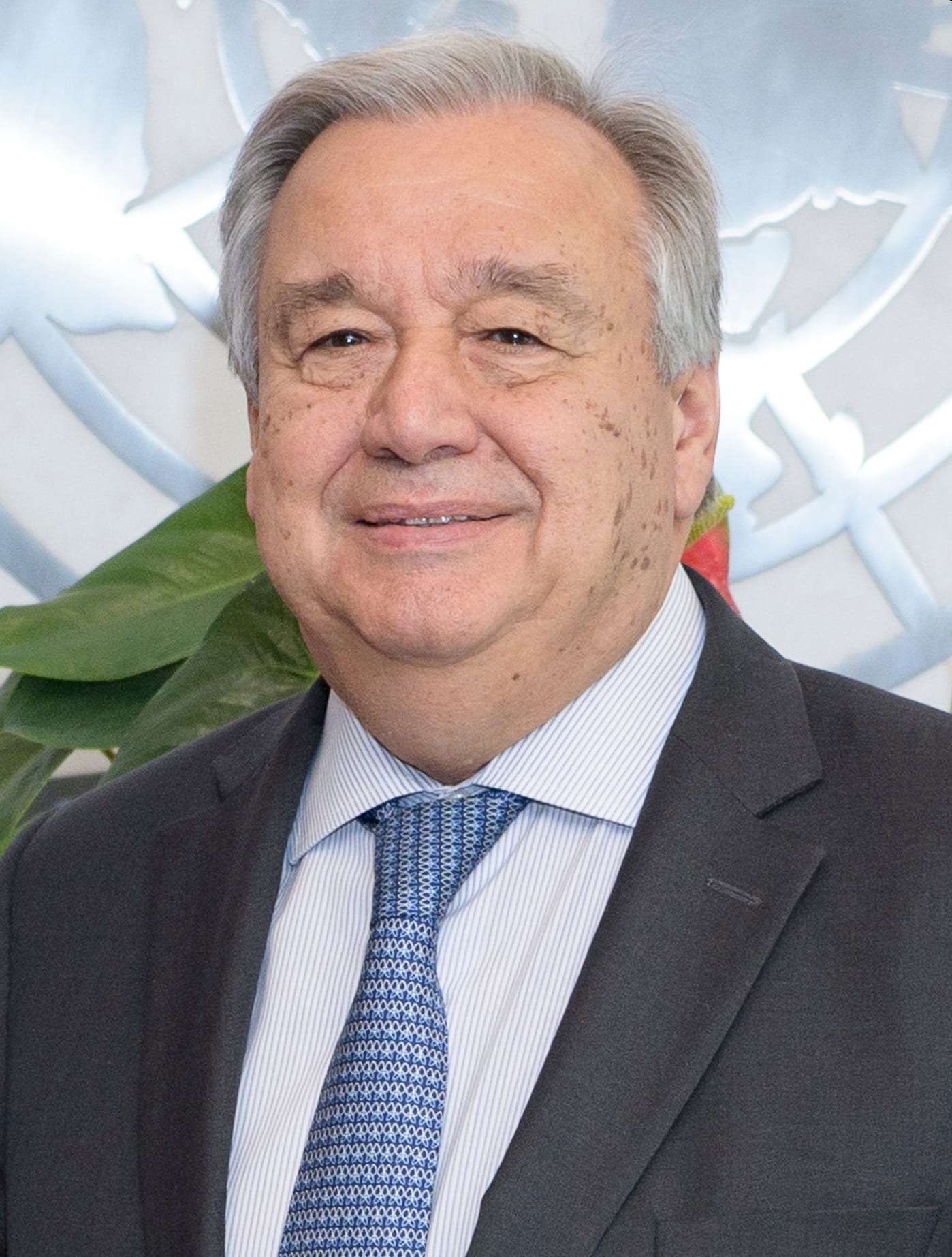 Un hombre mayor de traje oscuro y corbata azul posa sonriente ante el emblema de la ONU.