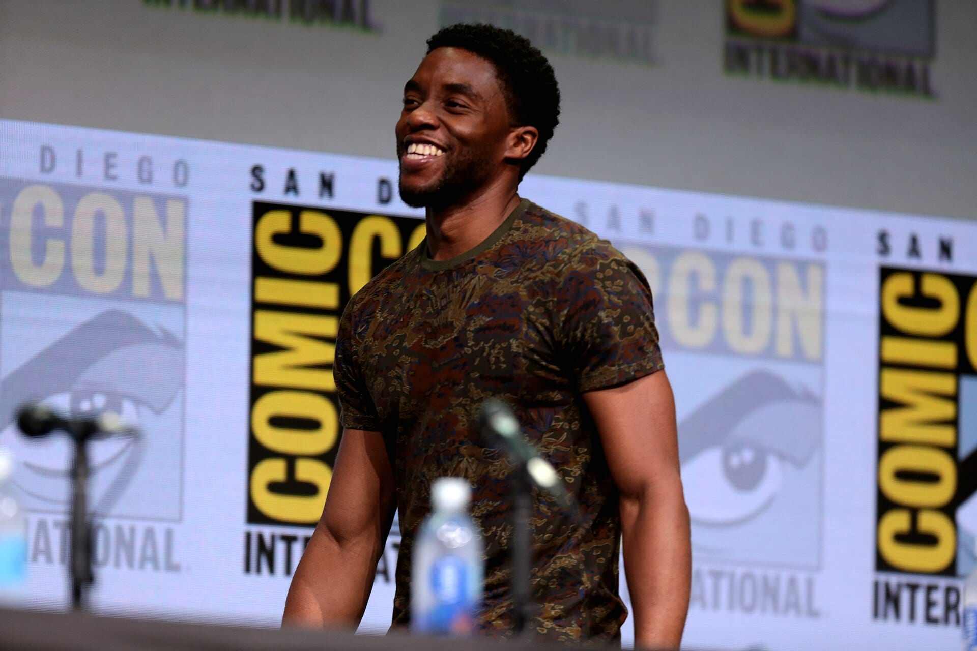 Chadwick Boseman sonríe de pie en un panel de Comic-Con, con micrófonos al frente y el logotipo del evento detrás.
