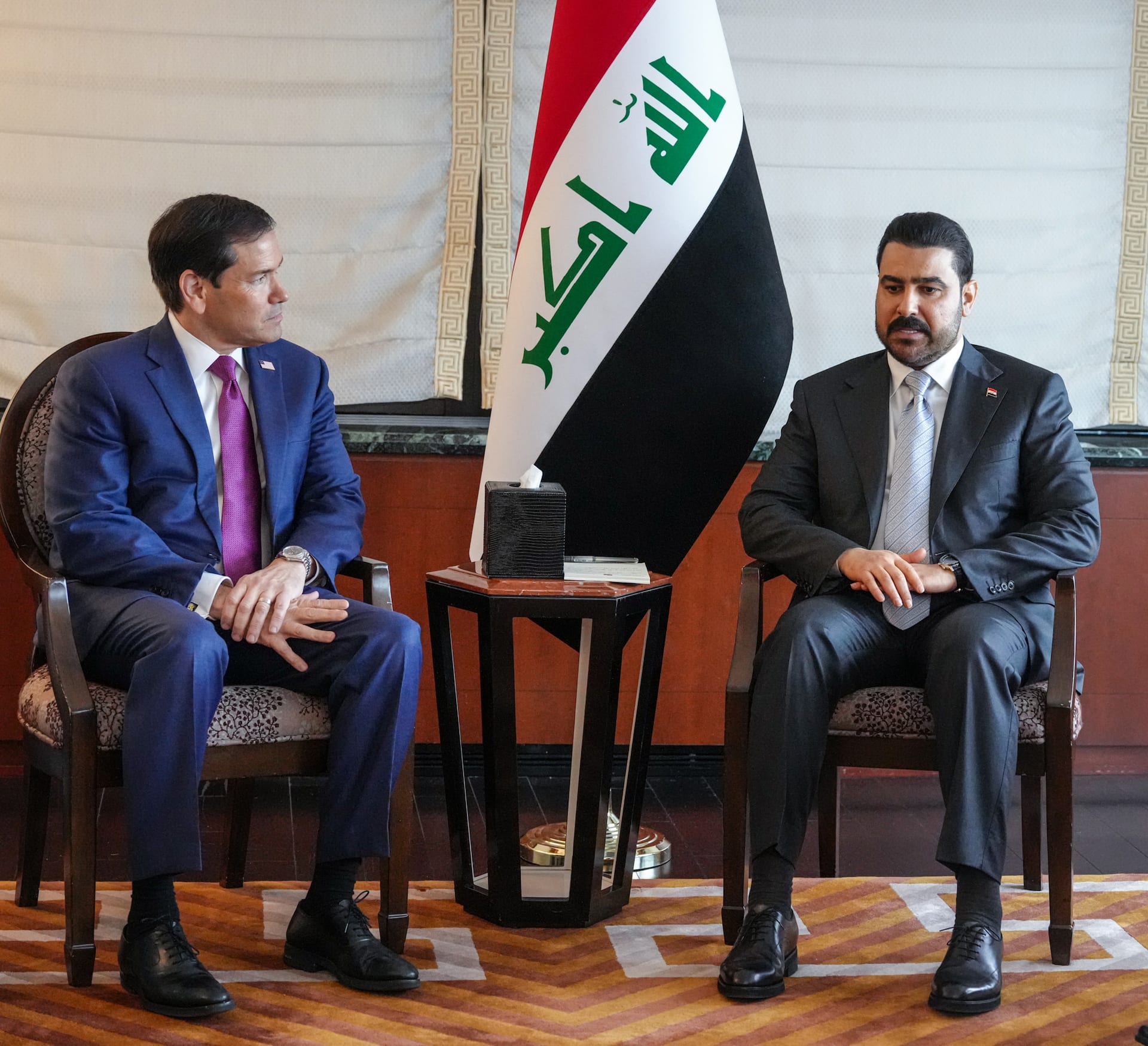 Marco Rubio y el primer ministro iraquí Ali al Zaidi conversan sentados en sillones junto a una bandera de Irak.