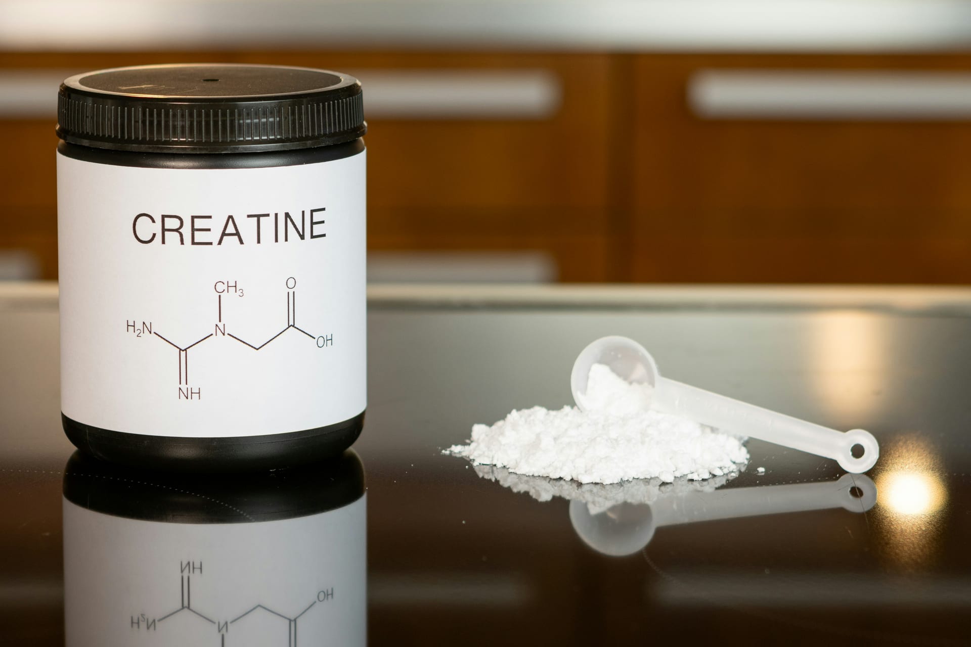 Un bote negro de creatina con su fórmula química impresa, junto a un montón de polvo blanco y una cuchara medidora sobre una superficie reflejante.