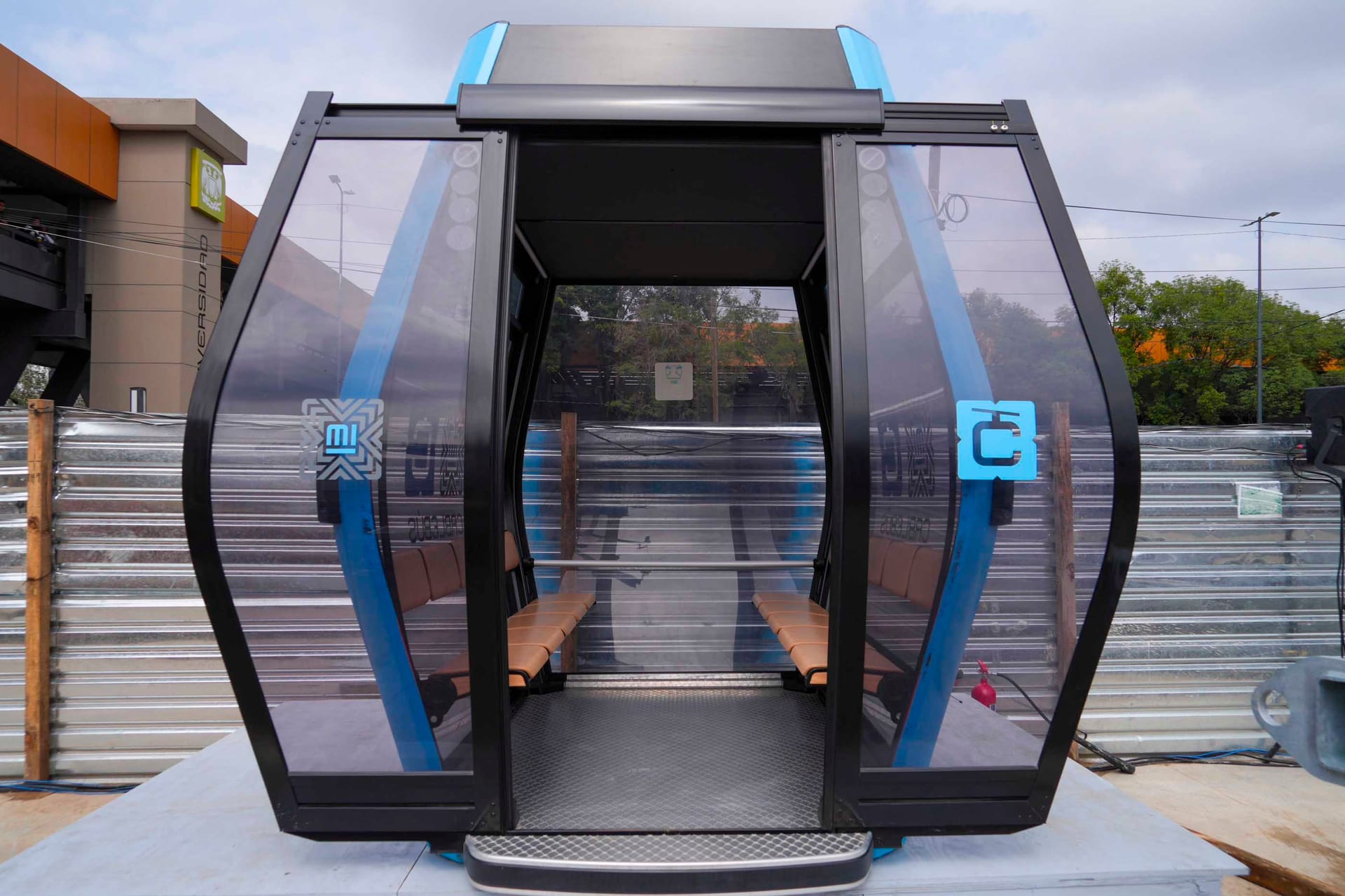 Una cabina vacía del Cablebús, con las puertas abiertas, frente a un edificio con un letrero que dice "Universidad".
