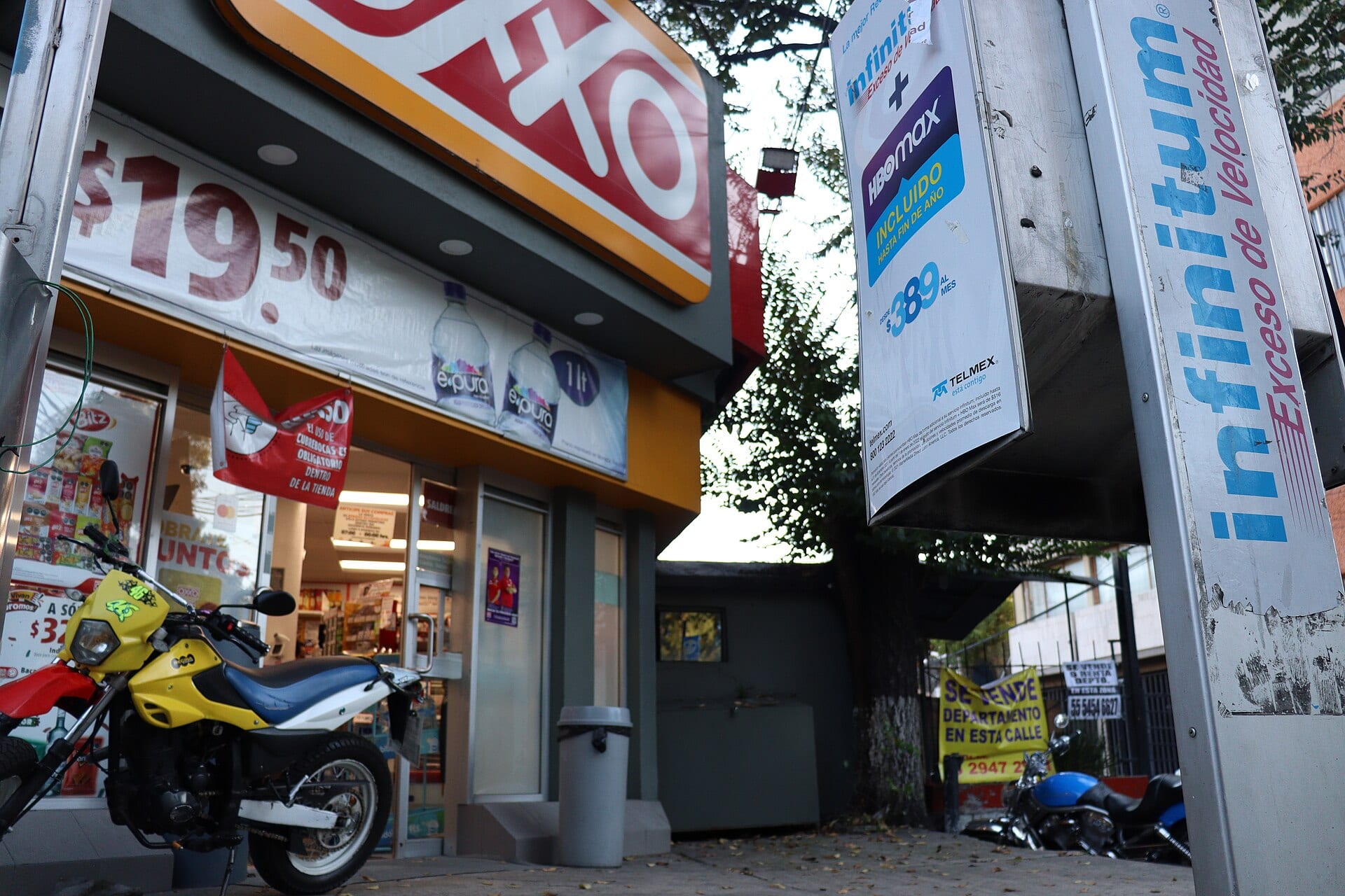 Fachada de una tienda de conveniencia Oxxo con su letrero rojo iluminado y anuncios promocionales en la entrada.