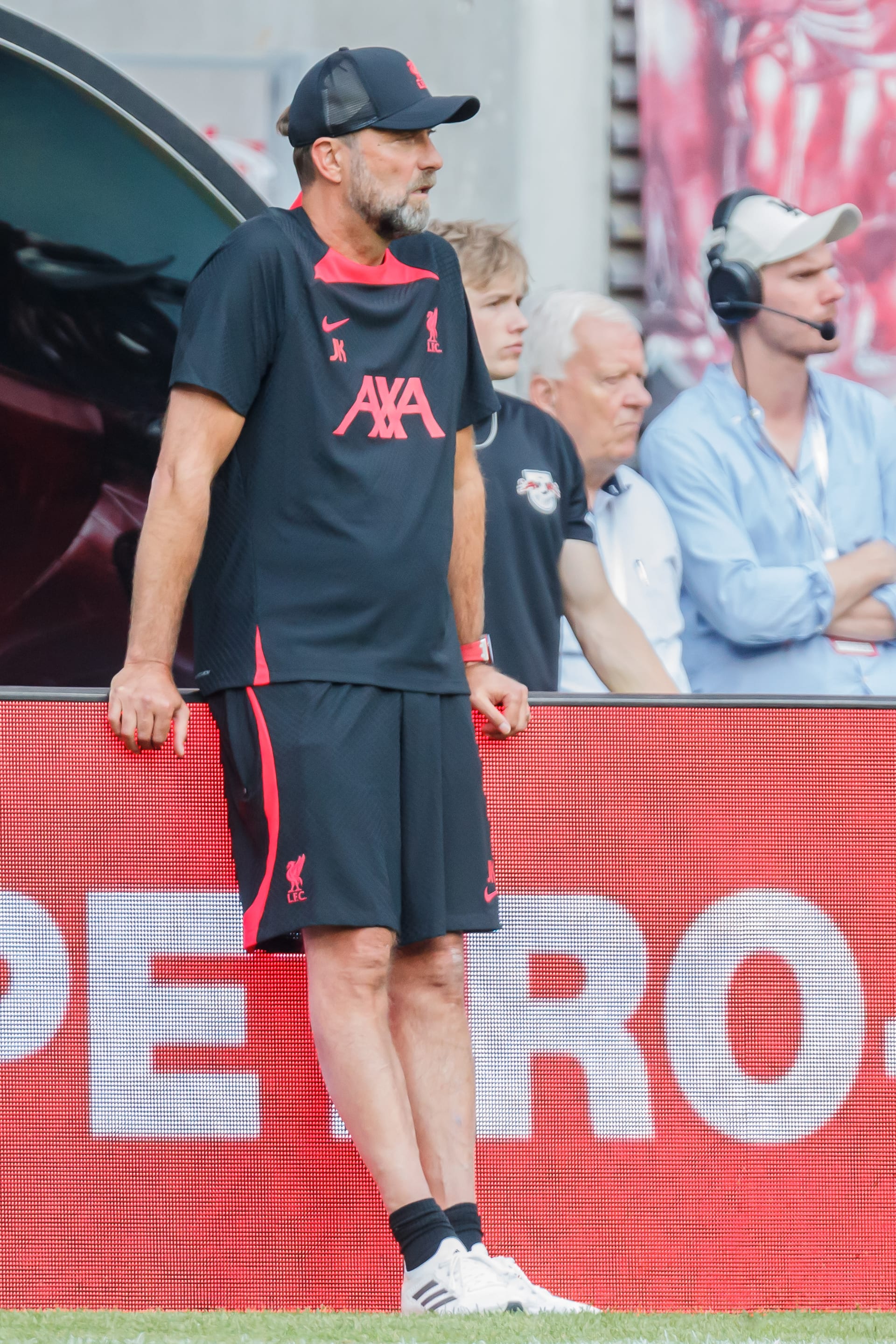 Un entrenador con gorra y chamarra oscura, con barba, gesticula desde la línea de banda durante un partido.