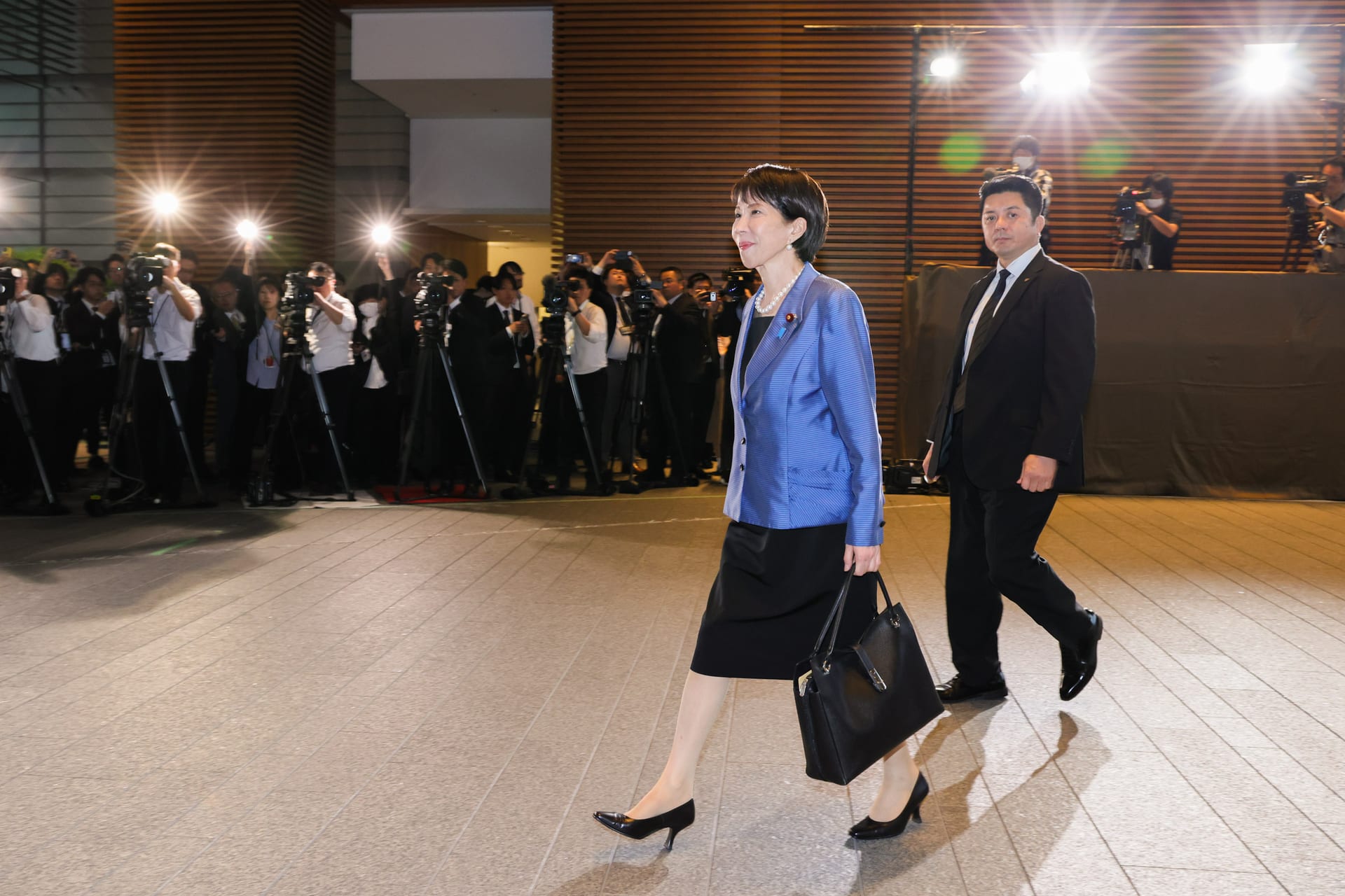 Sanae Takaichi camina por un pasillo de paredes de madera clara, vestida con un traje formal oscuro.