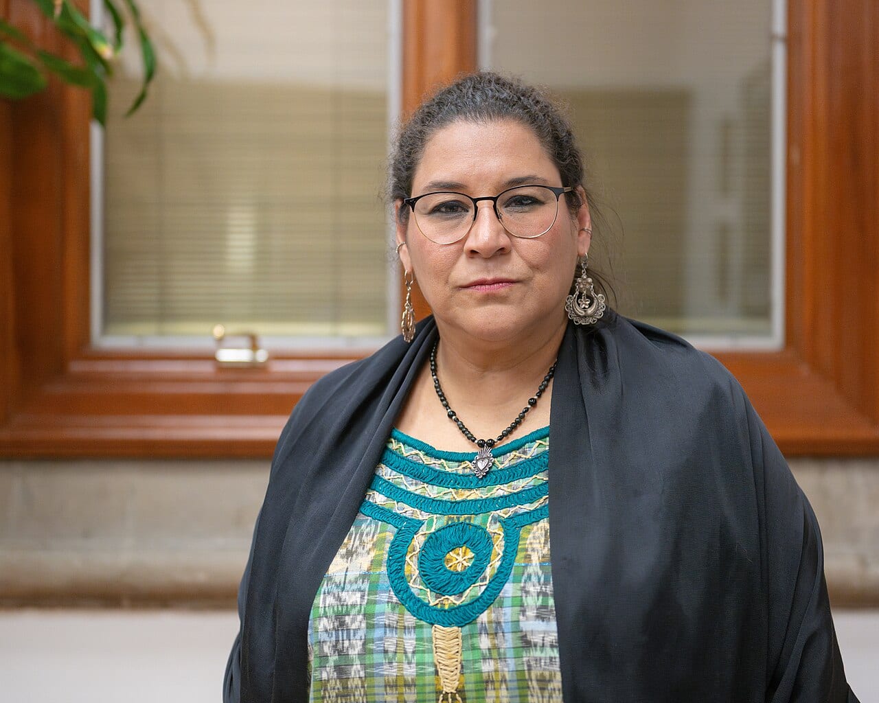 Una mujer con lentes y cabello oscuro recogido, envuelta en un chal negro sobre una blusa bordada en verde y turquesa.