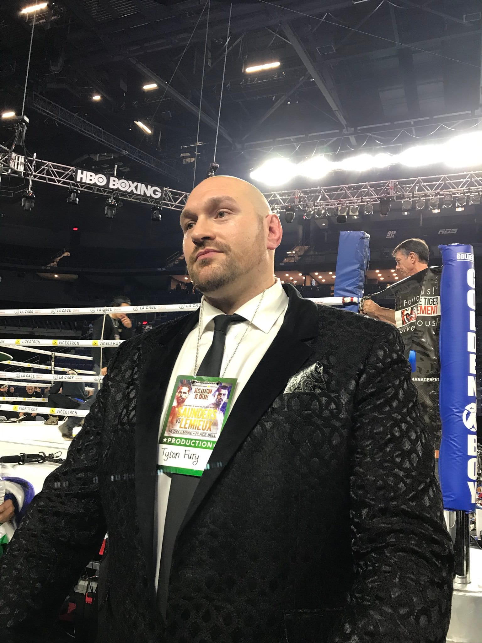 Tyson Fury, de traje negro y con un gafete de acreditación al cuello, de pie junto al ring en un evento de boxeo.