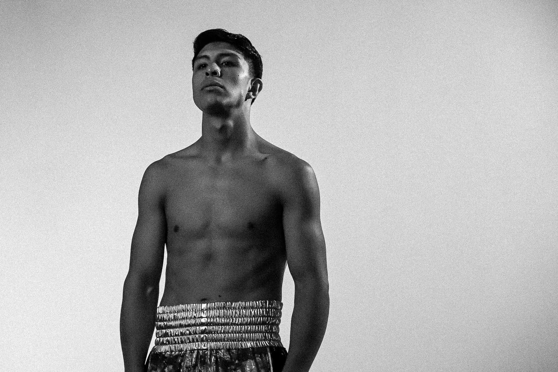 Fotografía en blanco y negro de un boxeador con el torso descubierto y el short de cintura metálica, de pie con gesto serio.