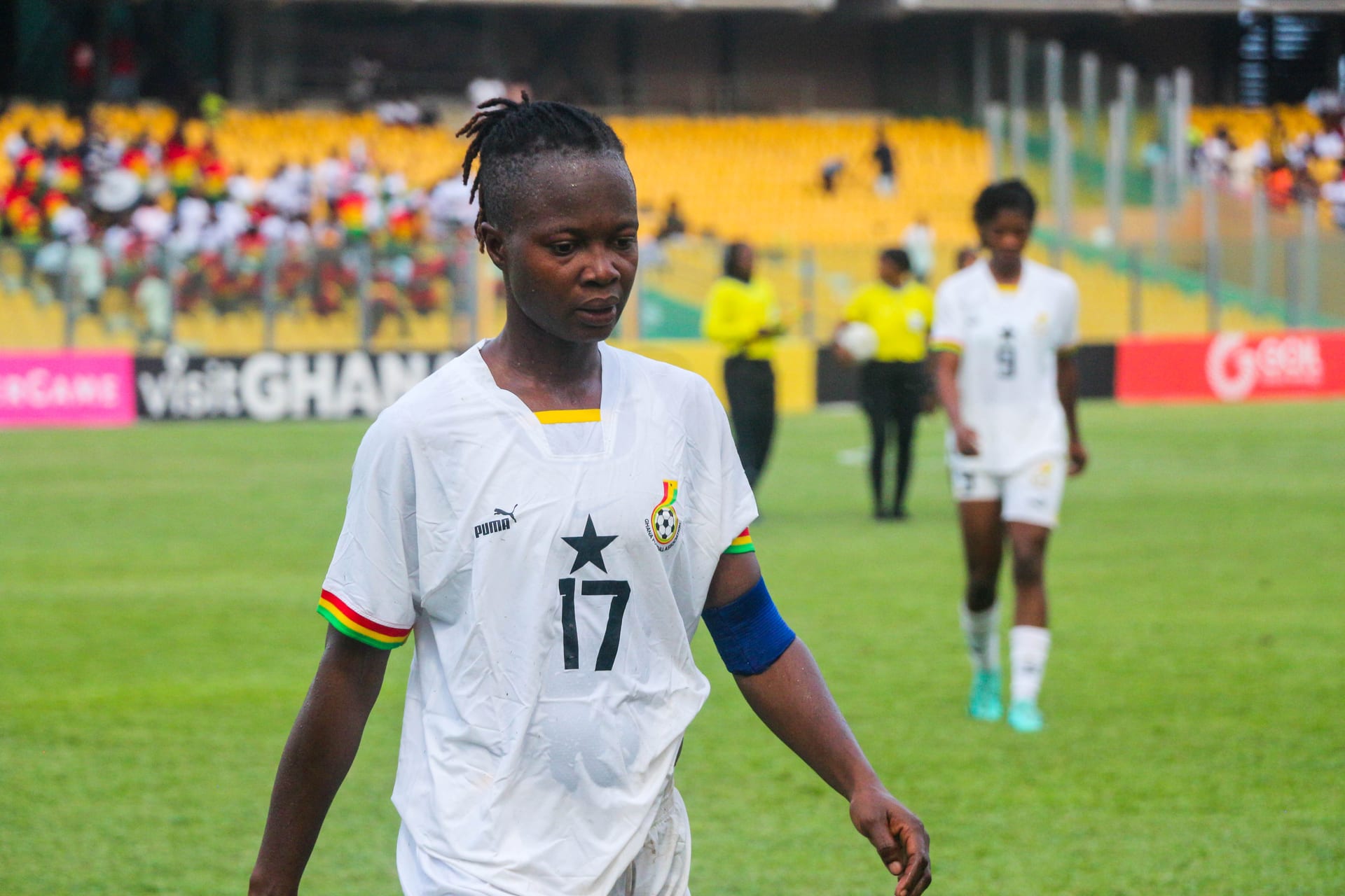 Una futbolista con camiseta blanca de Ghana y el número 17 porta un brazalete azul de capitana mientras camina por la cancha de un estadio con gradas amarillas, en un partido con su selección.