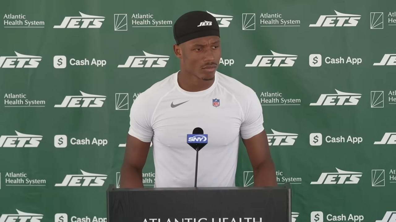 Un jugador con gorra y playera blanca de los Jets habla ante un micrófono en un podio, con un fondo de logos del equipo.