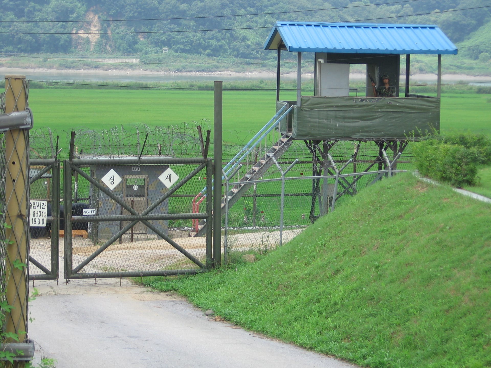 Un puesto de vigilancia elevado con techo azul junto a una valla de alambre de púas y un portón metálico, en un paisaje rural verde.