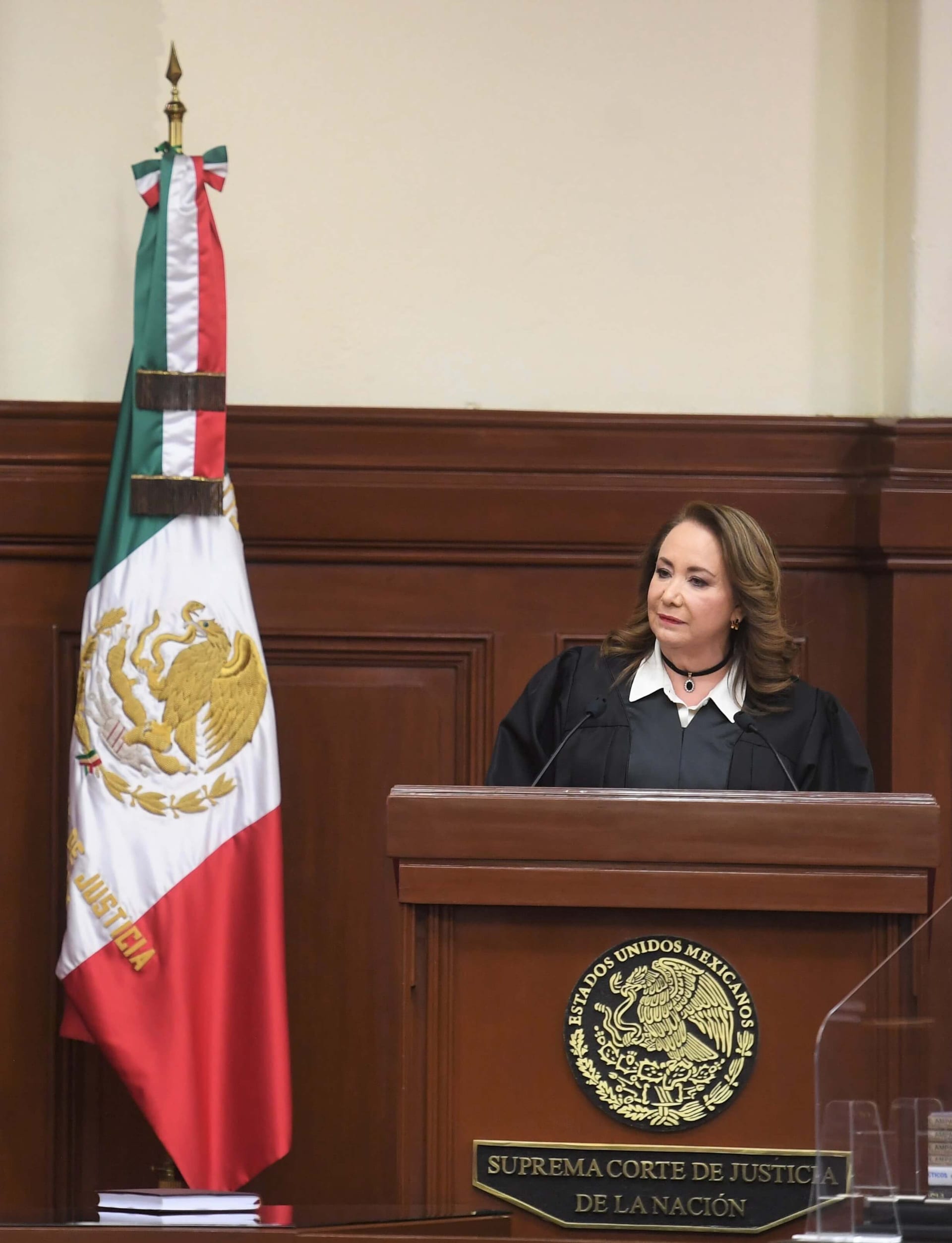 La ministra Yasmín Esquivel, con toga judicial, durante una sesión del Pleno de la Suprema Corte, con la bandera mexicana de fondo.