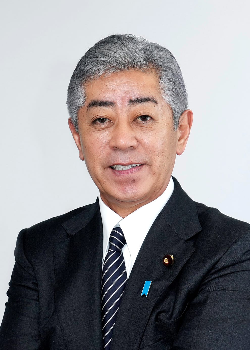 Retrato formal de Takeshi Iwaya con traje oscuro y corbata a rayas, sobre fondo gris liso.