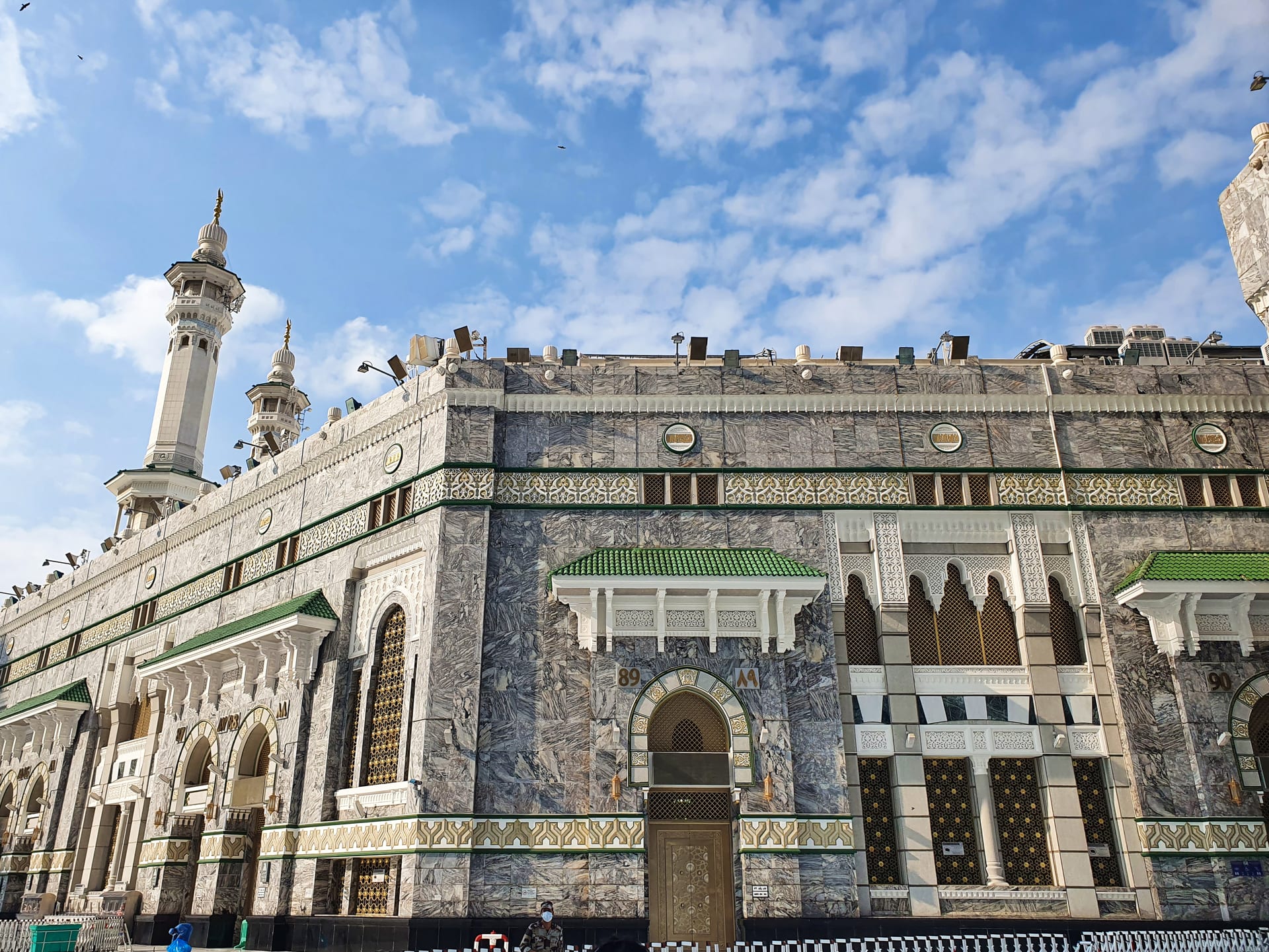 Fachada de mármol tallado de la Gran Mezquita de La Meca, con un minarete y puertas ornamentadas bajo un cielo con nubes dispersas.
